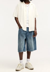 Man draagt een crèmekleurig overhemd met korte mouwen over een witte top, blauwe denim shorts tot op de knie, witte sokken en crèmekleurige sneakers met veters.