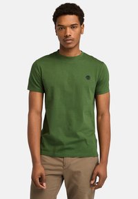 Ung mand iført en grøn t-shirt med korte ærmer og et lille sort logo på brystet, samt beige bukser, som står foran en ensfarvet baggrund.