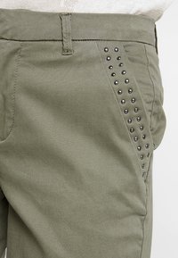 Olive zielone spodnie o gładkiej fakturze, z boczną kieszenią zdobioną czarnymi ćwiekami i prostym krojem.