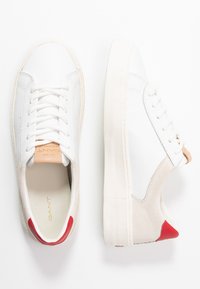 White leather sneakers with cream rubber sole and red accents. Features lace-up design, suede paneling, and embossed logo on insole.