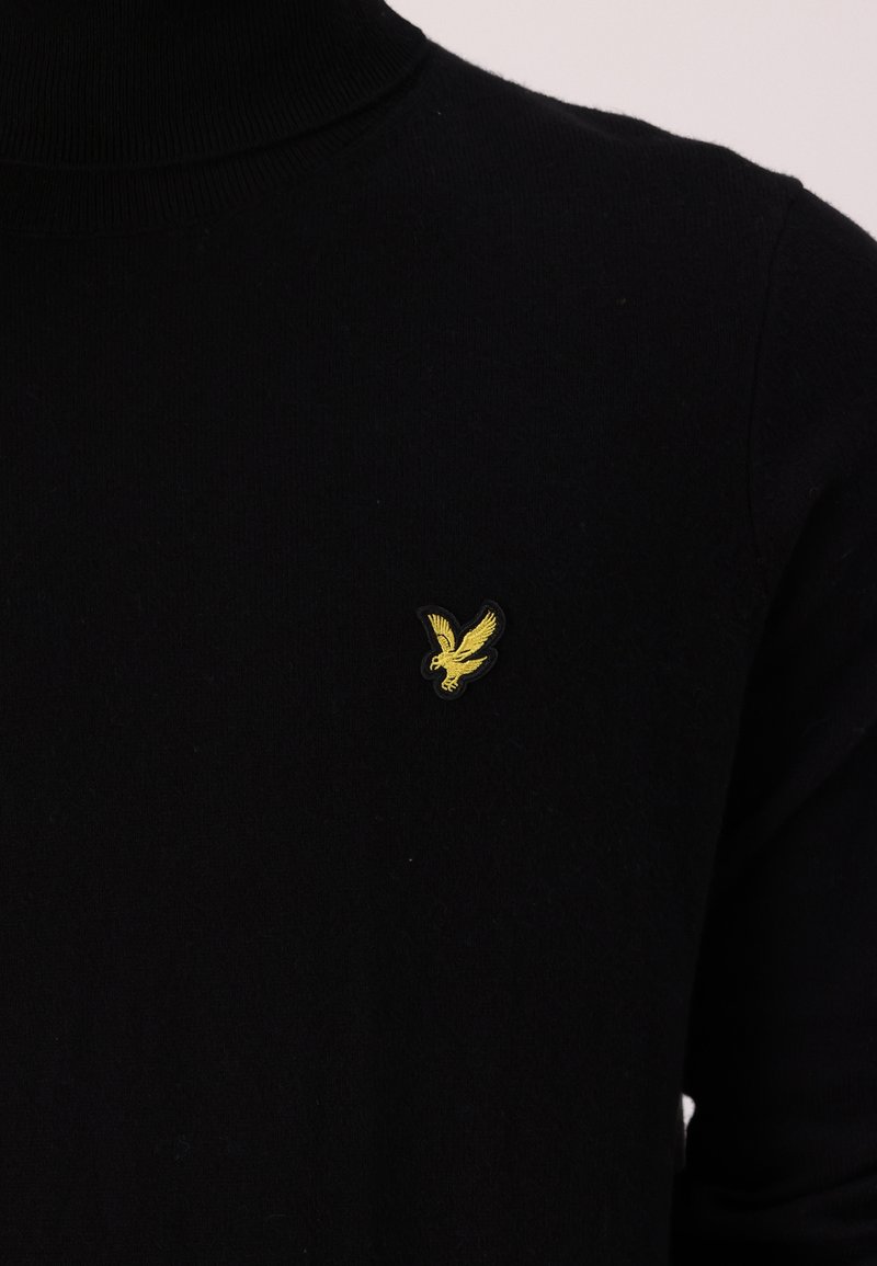 Pull à col roulé noir en tissu doux, avec un petit emblème d'aigle jaune sur la poitrine gauche. Design simple et ajusté.