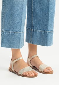 Płaskie sandały z metalicznymi złotymi paskami, otwarty nosek i zapięcie na kostkę. Noszone z przyciętymi dżinsowymi kulotami, prezentując swobodny wygląd.