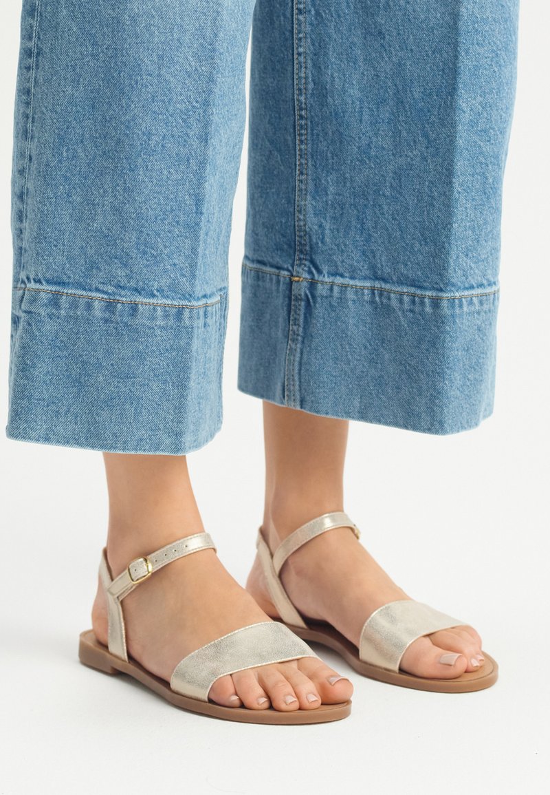 Płaskie sandały z metalicznymi złotymi paskami, otwarty nosek i zapięcie na kostkę. Noszone z przyciętymi dżinsowymi kulotami, prezentując swobodny wygląd.
