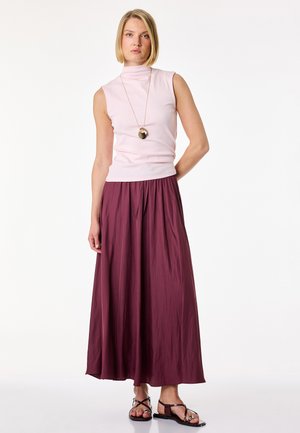 Donna in piedi che indossa un top rosa chiaro senza maniche, una lunga gonna bordeaux, una collana con ciondolo e sandali neri piatti su sfondo bianco.