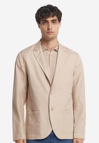 Ljusbeige linneblazer med enkelknäppt design, två framfickor och slaglappar, bärs över en beige polo-tröja.
