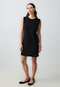Schwarzes ärmelloses Kleid mit lockerer Passform, Rundhalsausschnitt und seitlichen Schlitzen. Kombiniert mit schwarzen Ballerinas und subtilen goldenen Armreifen.
