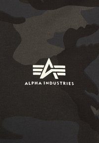 Camo-patterned fabric in shades of black and dark gray features a prominent white logo with "ALPHA INDUSTRIES" text.