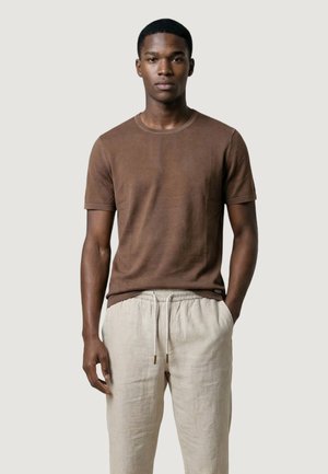 Giovane uomo in piedi, rivolto frontalmente, che indossa una camicia marrone a maniche corte e pantaloni beige con coulisse, su uno sfondo semplice.