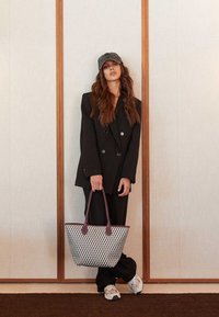 Torba na ramię z fakturowanym białym i szarym wzorem, fioletowymi uchwytami, noszona z czarnym oversize'owym żakietem i casualowymi trampkami.
