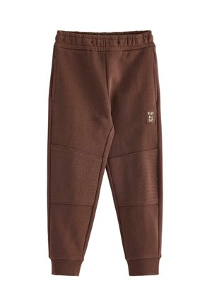 Junger Junge lächelt, trägt ein langärmeliges Shirt mit blauem Traktor-Design, braune Hose und hellblaue Turnschuhe mit roten Streifen.