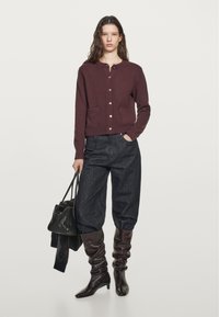 Burgundy knitted cardigan with round neckline, gold buttons, two front pockets, paired with dark denim baggy trousers and knee-high leather boots.
