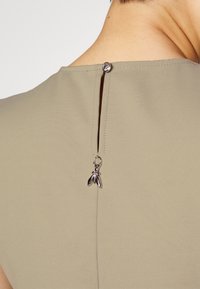 Gros plan sur l'encolure dos d'un vêtement beige avec un petit bouton en argent et un pendentif décoratif en argent suspendu sous l'ouverture en forme de trou de serrure.