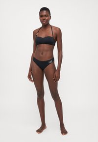 Crni bikini set koji se sastoji od bandeau topa i donjih dijelova niskog struka s detaljem loga. Glatka tkanina, minimalistički dizajn, pogodan za kupanje.