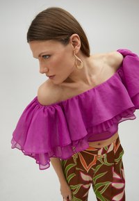 Lilla off-the-shoulder top med lagdelte flæser, parret med højtaljede, farverigt mønstrede bukser med bladmotiver.