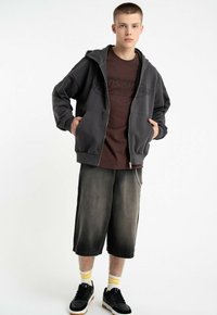 Szara bluza z kapturem na zamek, ciemno brązowa koszulka z grafiką, w zestawie z wyblakłymi czarnymi szortami. Czarne sneakersy z białymi podeszwami i żółte skarpetki w paski.