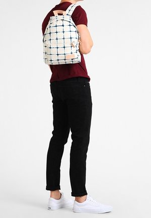 Osoba nosząca biały plecak w niebieską kratkę, bordo koszulę, czarne spodnie i białe trampki, stojąca na jednolitym tle.