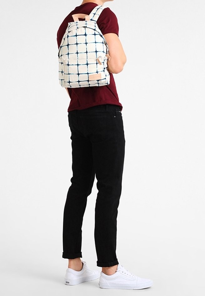 Personne portant un sac à dos blanc avec un motif à carreaux bleu, une chemise bordeaux, un pantalon noir et des baskets blanches, debout devant un fond uni.