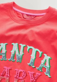 Červená tričko s krátkým rukávem a kulatým výstřihem, s vícebarevným vyšitým textem "SANTA" a "PARTY" v zelených a růžových akcent. 
