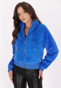 Giacca in pelliccia sintetica blu con chiusura a zip, collo alto e polsini a costine. Presenta due tasche con zip e un design corto.