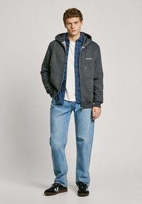 Sudadera gris con cremallera y cordones, superpuesta a una camisa de cuadros azul. Combinada con jeans azul claro y zapatillas negras.