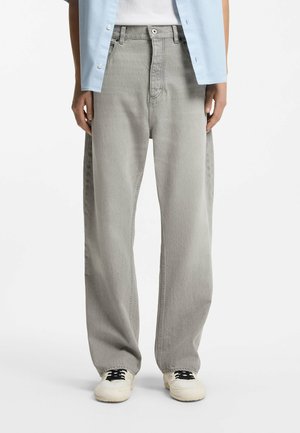 Person trägt hellblaues Hemd, weißes Top, lockere hellgraue Jeans und weiße Turnschuhe mit schwarzen Schnürsenkeln, steht vor weißem Hintergrund.