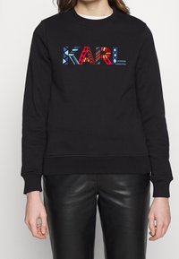 Černá mikina s nápisem "KARL" v barevných dekorativních písmenkách. Vyrobena z měkké tkaniny s vroubkovanými manžetami a lemem.