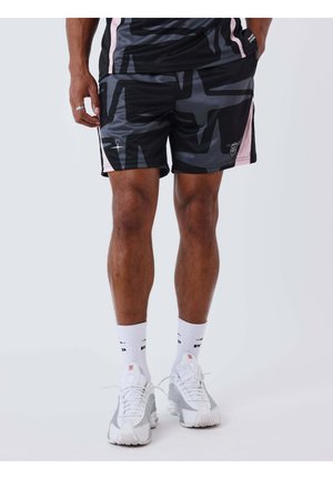 Man die zwarte en grijze korte broek met sterrenpatroon en roze accenten draagt, witte sportsokken en witte hardloopschoenen aan heeft, staand tegen een witte achtergrond.