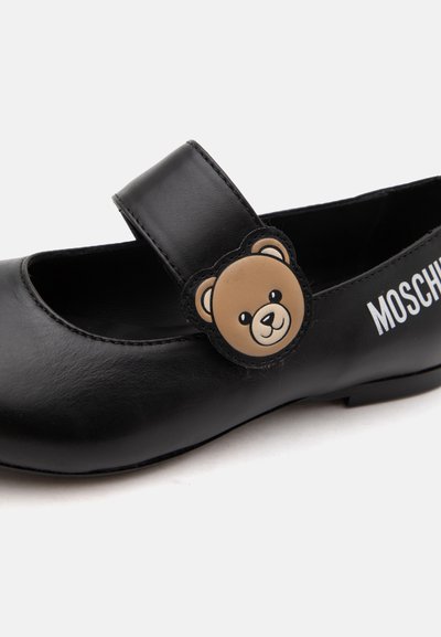 Chaussure Mary Jane noire avec une décoration visage d’ours sur la bride et le logo blanc Moschino sur le côté.