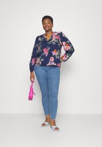 Blusa floral em azul com padrões em rosa e verde, decote em V, mangas longas; combinada com jeans skinny azuis e uma carteira rosa. Sandálias planas.