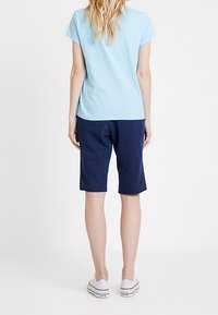 Frau steht mit dem Rücken zur Kamera, trägt ein hellblaues T-Shirt, marineblaue kniehohe Shorts und weiße Sneakers mit niedrigem Schaft.