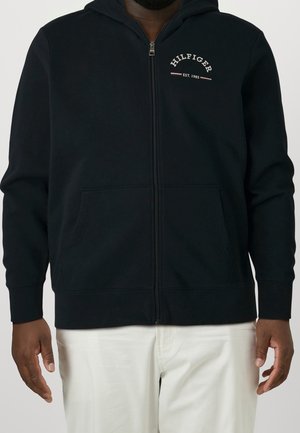 Mann trägt einen schwarzen Kapuzenpullover mit Reißverschluss mit dem Text "Hilfiger Est. 1985" auf der Brust und weiße Hosen vor einem einfarbigen Hintergrund.