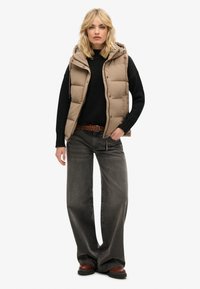 Beige wattierte Weste mit Kapuze über einem schwarzen Pullover, kombiniert mit grauen weit geschnittenen Jeans. Verfügt über einen geflochtenen Gürtel und dunkle braune Stiefel.