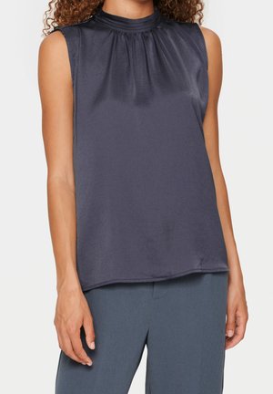 Bezrekawka, ciemnoszara satynowa bluzka z marszczonym wysokim kołnierzem noszona z niebieskimi, dopasowanymi spodniami na osobie z kręconymi włosami.