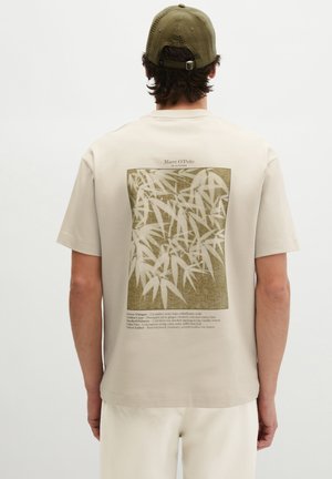 Mężczyzna w beżowej koszulce i oliwkowo zielonej czapce, plecami do kamery, na koszulce znajduje się prostokątny, abstrakcyjny wzór roślinny oraz tekst.