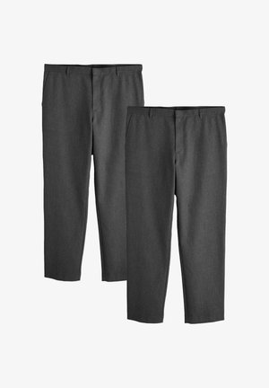 Férfi fonott hajjal, fekete pettyes dzsekit, világoskék inget, szürke nadrágot és fekete cipőt viselve, fehér háttér előtt áll.