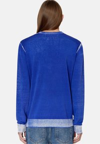 En person med långt brunt hår som bär en bleknad blå tröja med ljusblå muddar och fåll, tillsammans med blå denimjeans, står vänd bort.