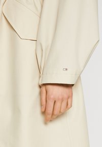 Trench coat impermeabile beige con una vestibilità rilassata. Presenta una grande tasca, dettagli sui polsini e un piccolo logo in metallo sull manica.