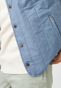 Hellblaues gesteppte Jacke mit braunen Druckknöpfen und einem seitlichen Taschenlabel. Getragen über einem weißen Hoodie und grünen Hosen. Strukturiertes Material.