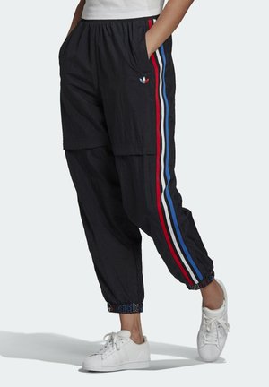 Person iført sorte Adidas træningsbukser med røde, hvide og blå sidelommer samt hvide sneakers, hænderne i lommerne, står på ét ben.