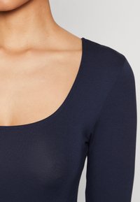 Mörkblå topp med långa ärmar, gjord av ribbat tyg. Har en bred rund halsringning och figursydd siluett. Slät textur med minimala sömmar.