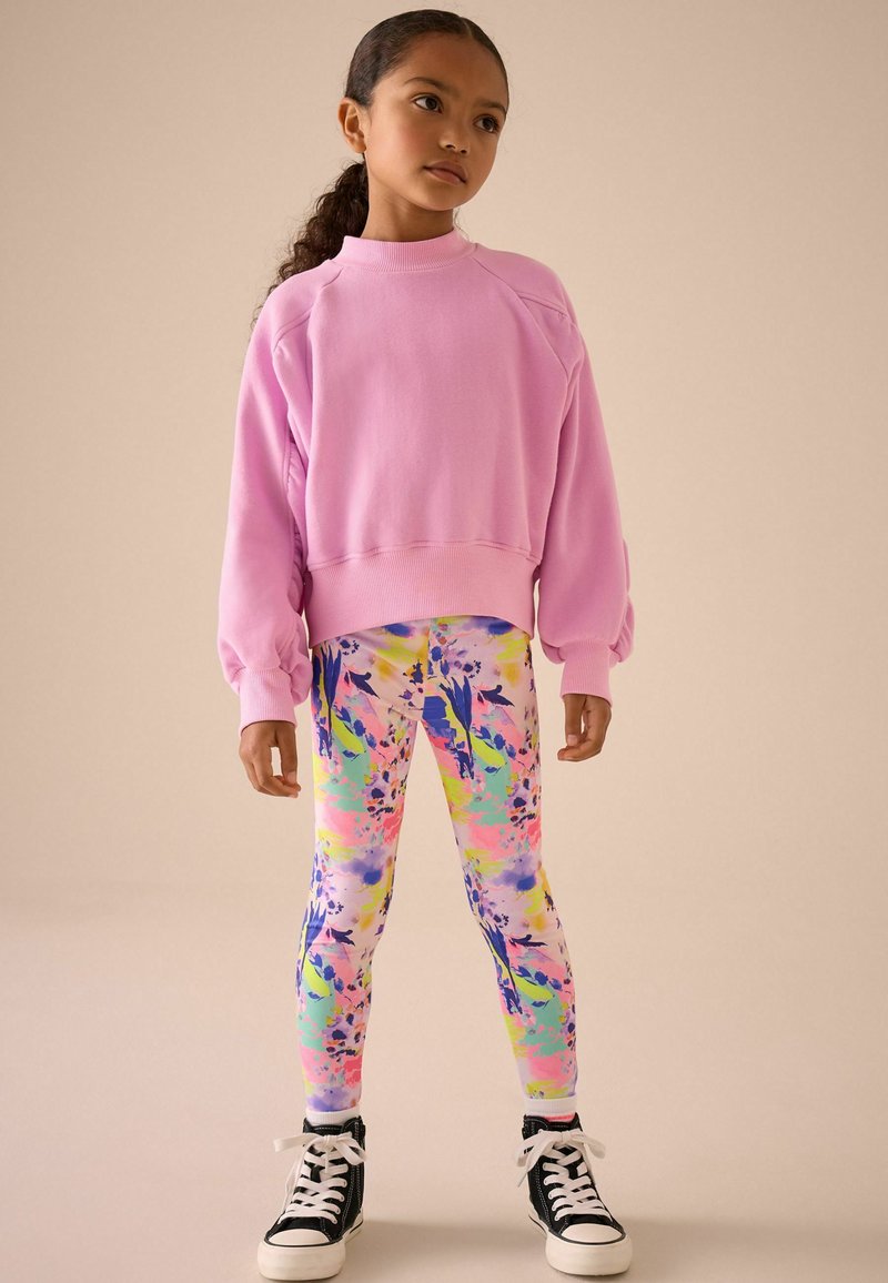 Rózsaszín pulóver puffos ujjakkal, mult színű virágmintás leggings-szel párosítva. Fekete felsőrésszel és fehér talppal rendelkező sportcipő. Lazább, színes viselet.