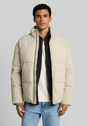 Young man wearing a beige puffer jacket over a black button shirt and white t-shirt, paired with light blue jeans, standing against gray background.