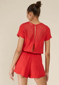 Combinaison rouge avec un tissu texturé. Manches courtes, un dos à motif en losanges et un détail en forme de trou de serrure au niveau du cou. Taille élastique et shorts.