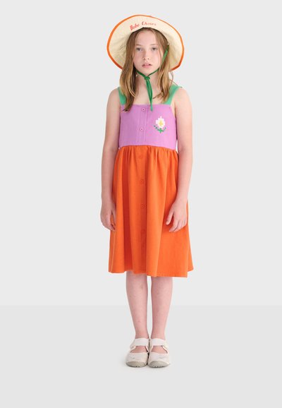 Jeune fille portant une robe sans manches violette et orange avec un détail floral et un chapeau à large bord avec des brides vertes sous le menton, debout devant un fond blanc.
