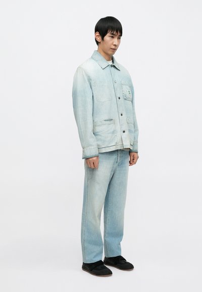 Φωτεινό γαλάζιο τζιν μπουφάν με μυτερό γιακά και δύο μπροστινές τσέπες, συνδυασμένο με παντελόνι από τζιν στο ίδιο φωτεινό γαλάζιο χρώμα και μαύρα παπούτσια.