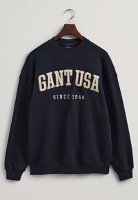 Námořnická modrá mikina s kulatým výstřihem, na které vyniká výrazný nápis "GANT USA" a "SINCE 1949" v bílé a zlaté barvě. Měkká tkanina, žebrované manžety.