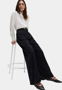 Camisa blanca de botones con mangas largas, pantalones negros de cintura alta y de pierna ancha, y zapatos planos negros con cordones, sentada en un taburete blanco.