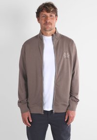 Sweat-shirt zippé marron avec col haut, surpiqûres blanches et un petit motif brodé sur la poitrine gauche. T-shirt blanc en dessous.