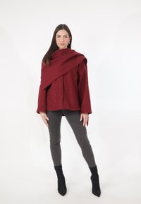 Cappotto bordeaux testurizzato con collo a scialle asimmetrico. Presenta maniche ampie, davanti aperto, indossato sopra jeans skinny grigi e stivaletti neri.