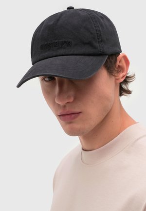 Hombre joven con una gorra negra con bordado sutil y una camiseta de cuello redondo beige claro, frente a un fondo liso.